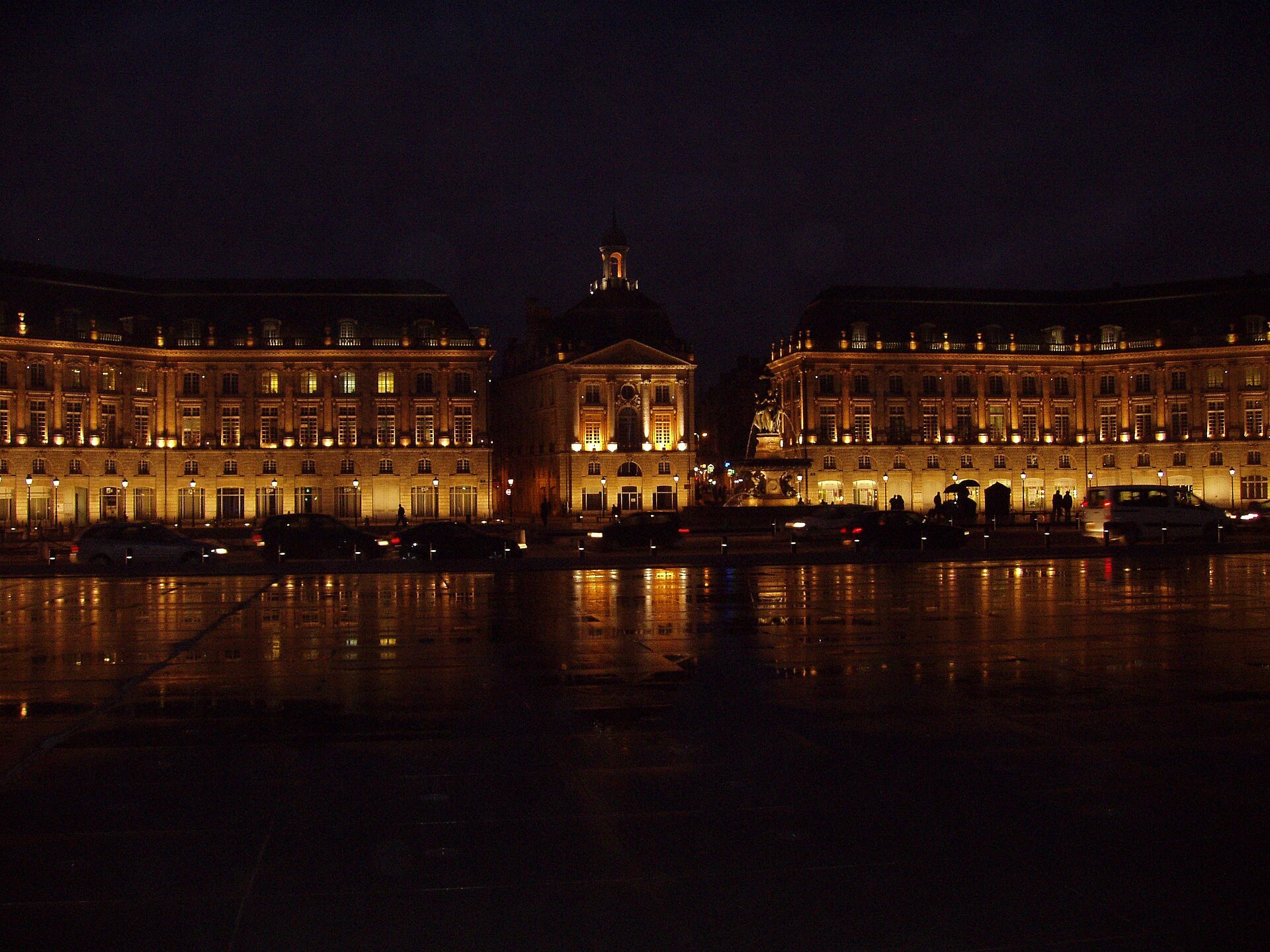 Place de la Bourse de Bordeaux de nuit, pavé mouillé, façades éclairées et fontaine des Trois Grâces.