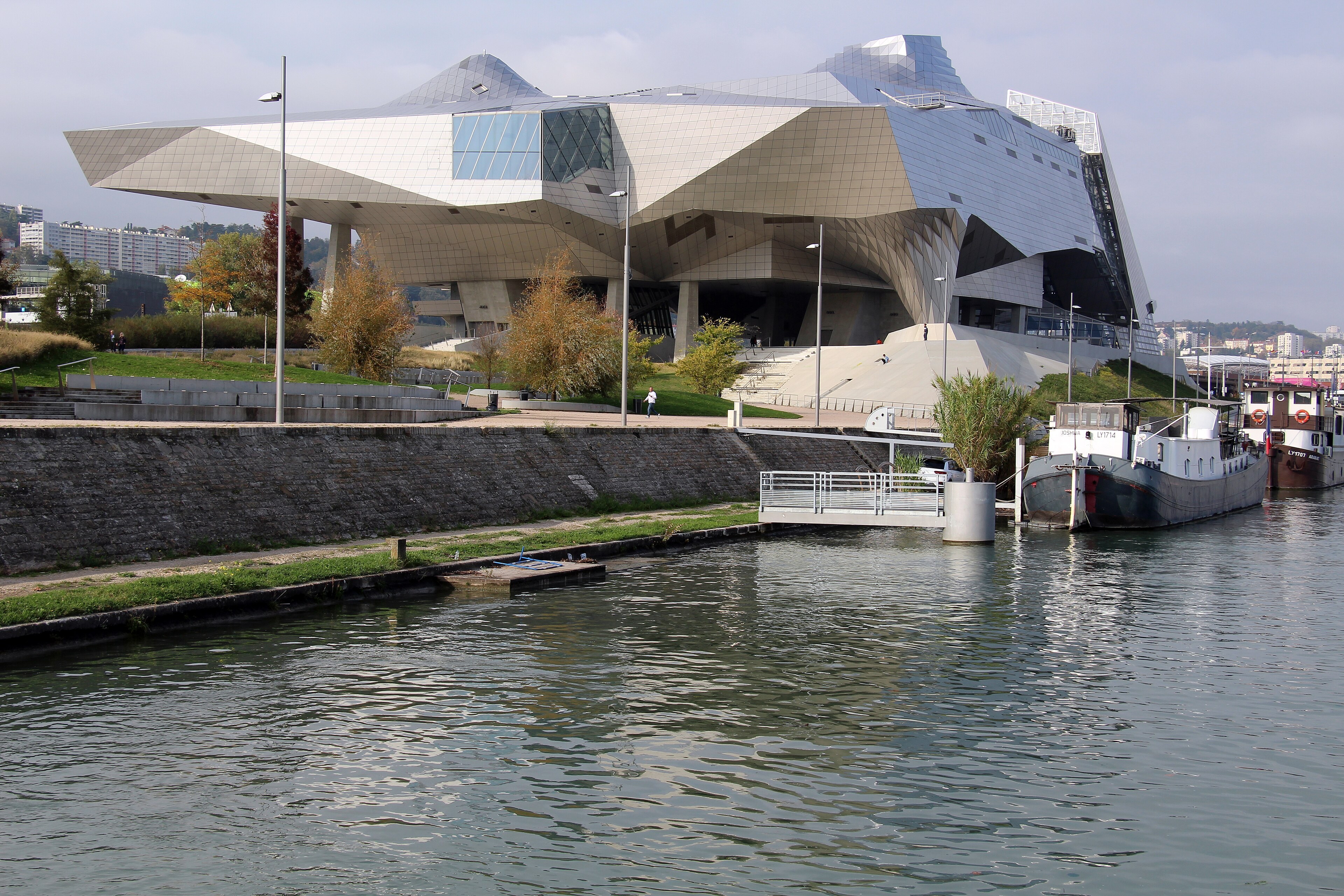 Musée des Confluences vu depuis le quai : volumes de verre, berges et bateaux à quai.