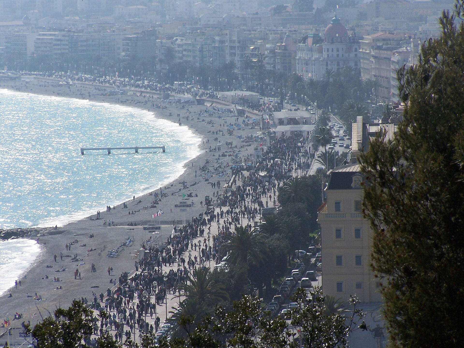 Promenade des Anglais vue en hauteur : plage de galets, foule sur le trottoir, dôme du Negresco au loin.